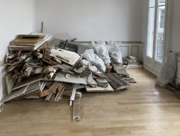 Room with pile of demolition debris including wood, doors, and sink. White walls and wood floor.