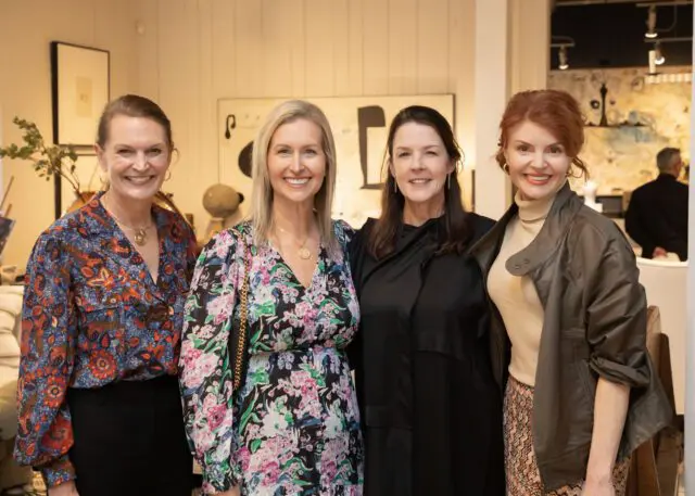 Four women smiling, two in floral dresses, one in a patterned blouse, one in a black dress and