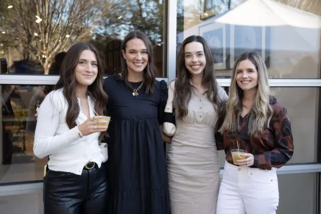 Four women smiling, holding drinks, in front of a building with large windows.