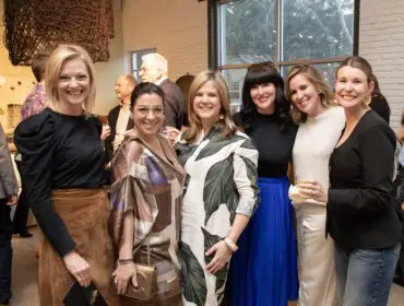 Six women smiling at a gathering. One woman wears a black top and brown skirt.