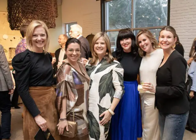 Six women smiling at a gathering. One woman wears a black top and brown skirt.