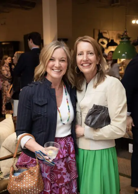 Two women smiling, one in a navy pinstripe jacket and floral skirt, the other in a cream jacket and