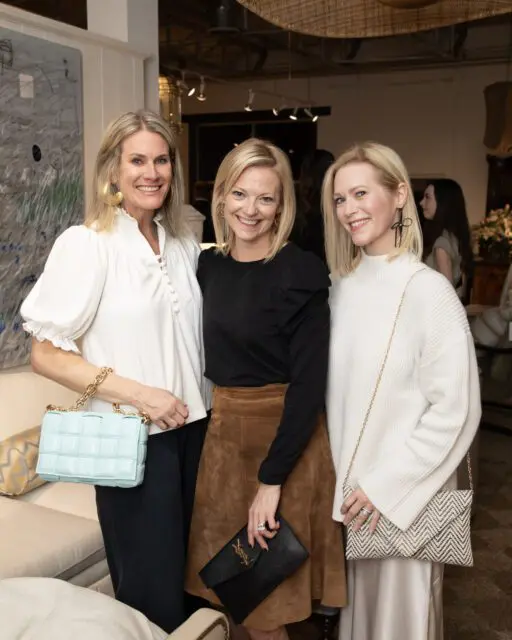Three women smiling, one in white with a light blue bag, one in black with a brown skirt and black