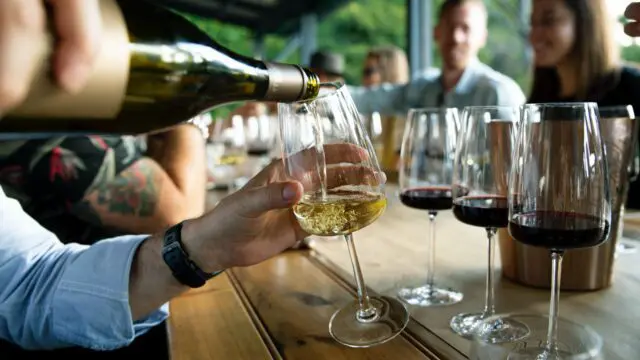 White wine being poured into a glass with red wine in glasses in the background.