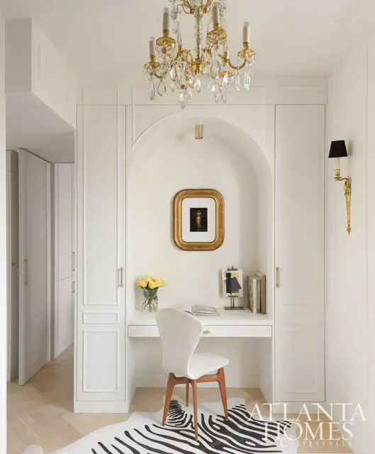White desk with chair, gold framed art, and zebra rug under a crystal chandelier.