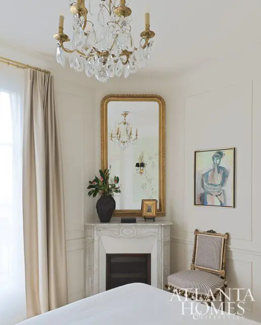 Bedroom with marble fireplace, gold mirror, crystal chandelier, and striped chair.