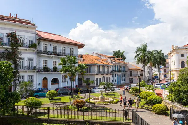 Light blue buildings with red tile roofs, balconies, and palm trees line a street with a fountain