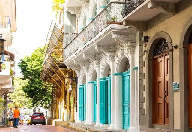 White building with ornate columns and turquoise doors, next to a tan building with brown doors.