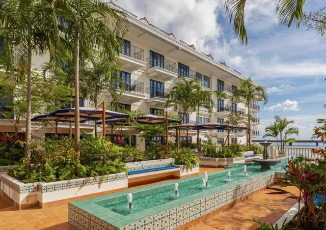 White hotel with balconies, palm trees, and a long water fountain feature.