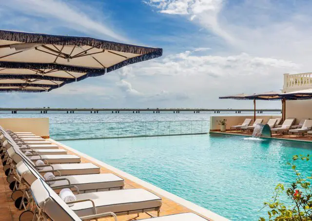 Infinity pool with lounge chairs and umbrellas overlooking a body of water with a bridge in the