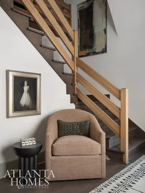 Tan armchair with patterned pillow next to a dark side table and a wooden staircase.