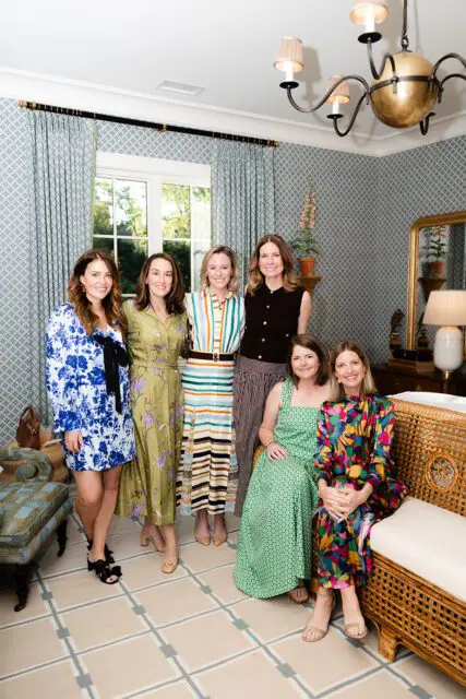 Six women posing in a room with blue patterned wallpaper, patterned curtains, and a rattan sofa.