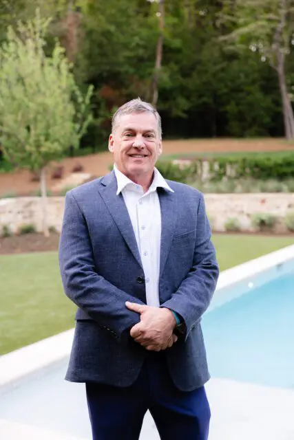 Man in blue suit jacket and patterned shirt standing by a pool.