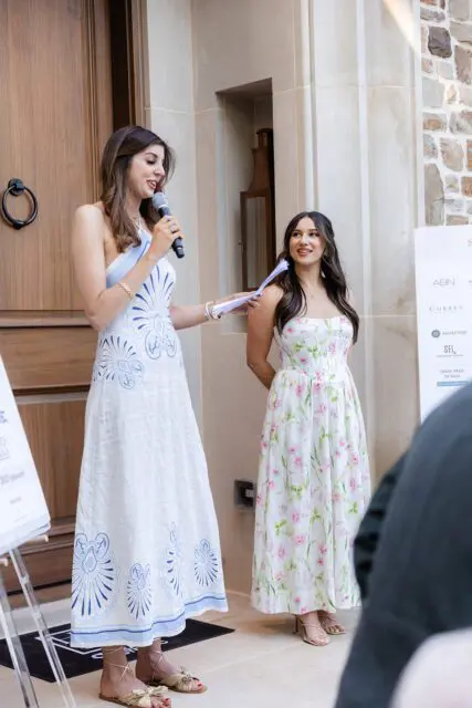 Woman in white dress with blue pattern speaks into microphone, woman in floral dress stands beside