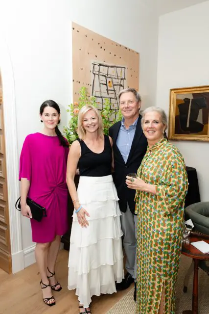 Four people posing indoors. A woman in a pink dress, a woman in a black top and white skirt, a man