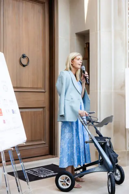 Woman in blue floral dress and light blue blazer speaks into a microphone