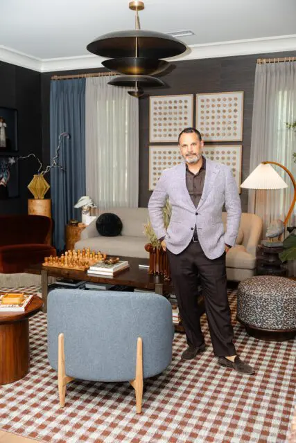 Man in blazer standing in living room with checkered rug, chess set, and modern light fixture.