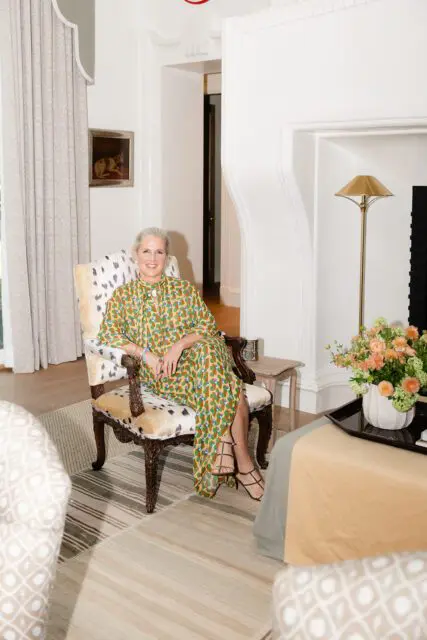 Woman in patterned dress sitting in upholstered chair with fireplace and flowers in background