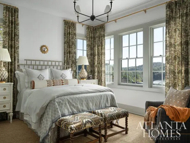 Bedroom with a grey metal bed, patterned curtains, and three windows overlooking a landscape.