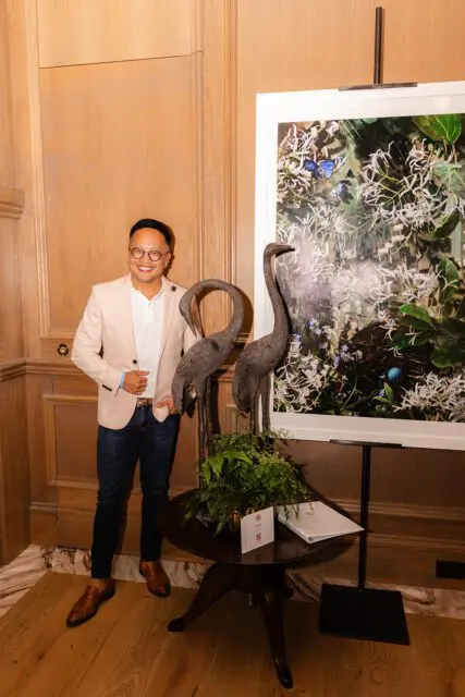 Man in light pink blazer and dark jeans standing next to artwork and bird sculptures.