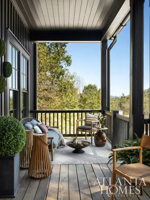 Covered porch with dark wood siding, seating area with chairs, sofa, fire pit, and view of trees.