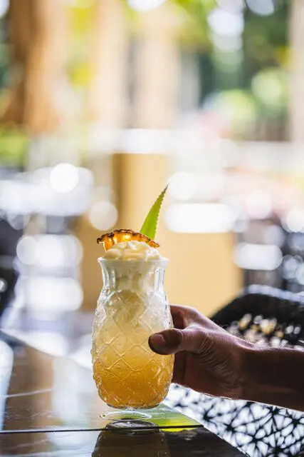 Hand holding a pineapple-shaped glass filled with a frothy yellow drink, garnished with pineapple