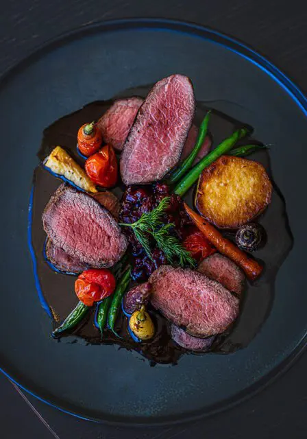 Slices of rare venison with roasted vegetables and berry compote on a dark plate