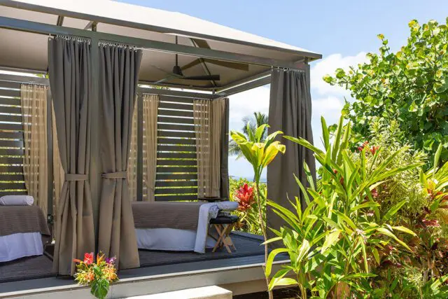 Outdoor spa cabana with massage tables, gray curtains, and lush tropical foliage.
