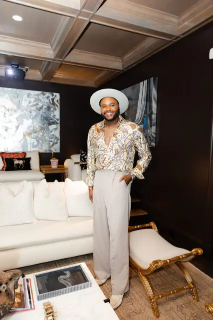 Man in white hat and patterned shirt stands in a living room with white sofa and coffered ceiling.