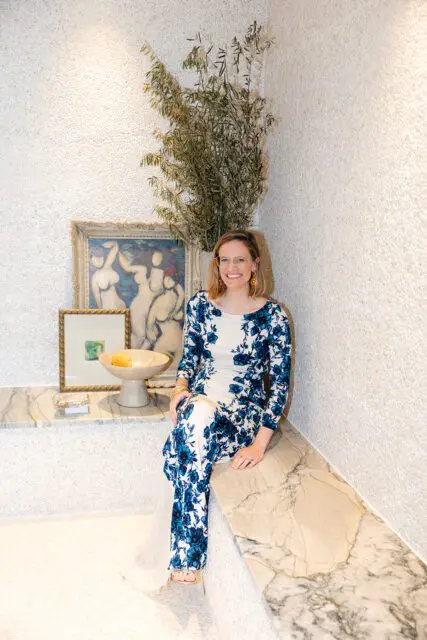 Woman in blue floral dress sitting on marble bench, with artwork and plants behind her.