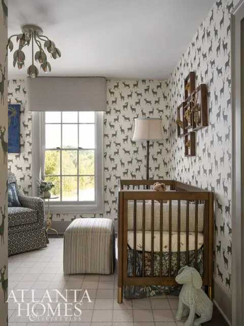 Nursery with deer wallpaper, wooden crib, striped ottoman, and patterned armchair.