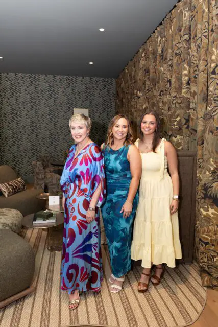 Three women posing in a room with patterned wallpaper and floral curtains.
