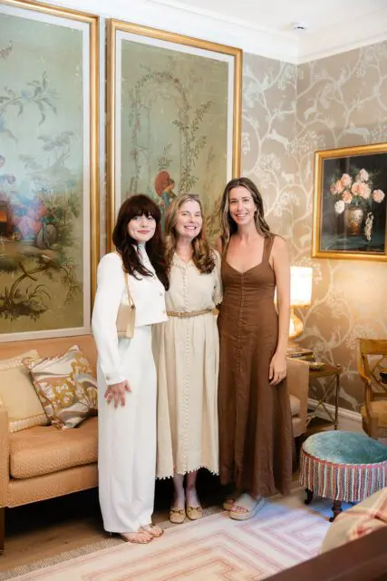 Three women in a room with ornate wallpaper and framed art. One wears white, one beige, one brown.