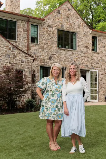 Two women standing on a lawn in front of a stone house.
