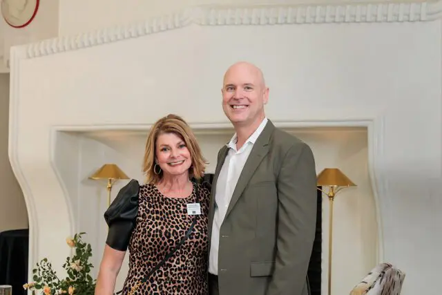 Woman in leopard print dress and man in olive suit smiling in front of fireplace