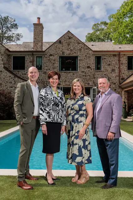 Four people standing in front of a stone house with a swimming pool