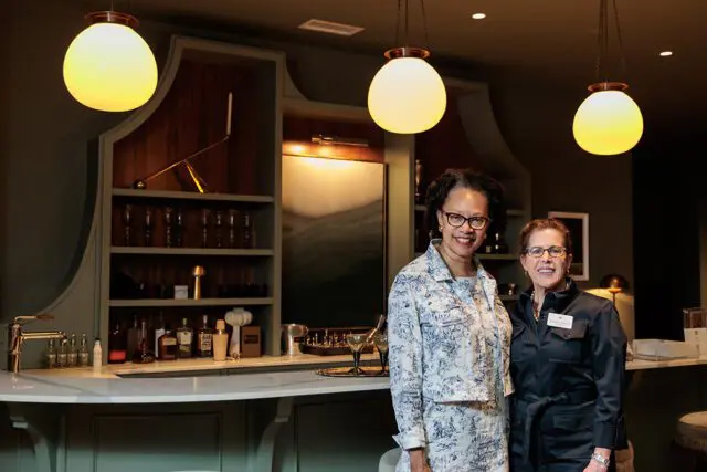 Two women stand in front of a bar with globe pendant lights and shelves stocked with bottles and