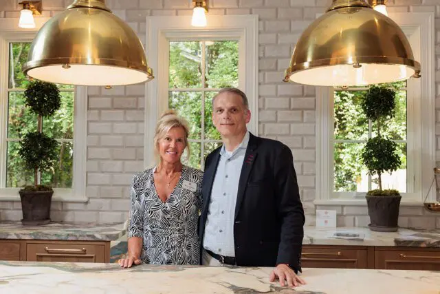 Woman in patterned dress and man in suit stand in kitchen with brass pendant lights and subway tile