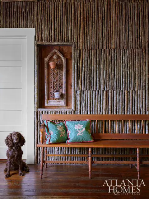 Wooden bench with floral pillows against a wall of vertical sticks. Dog sits nearby.