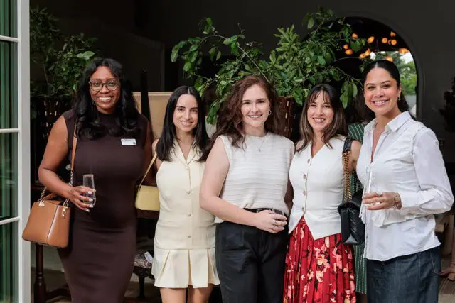 Five women smiling, some holding drinks, in an indoor setting with plants.
