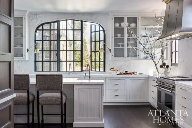 White kitchen with marble counters, arched window, and gray upholstered bar stools