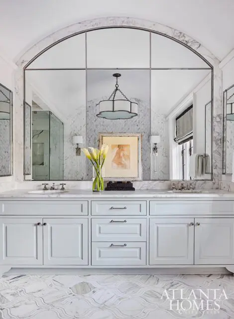 White bathroom vanity with marble countertop, arched mirror, and patterned marble floor.