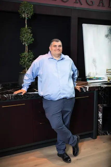 Man in blue dress shirt and gray pants leaning against a dark wood counter.