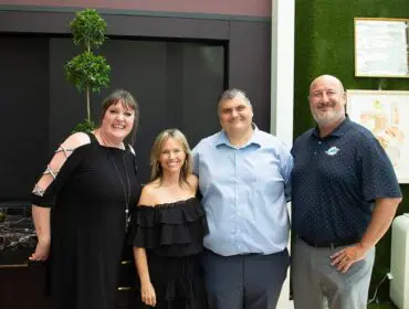Four people posing for a photo in front of a dark cabinet and a green wall with art.