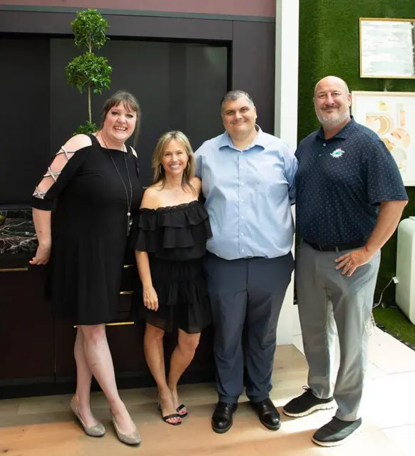 Four people posing for a photo in front of a dark cabinet and a green wall with art.
