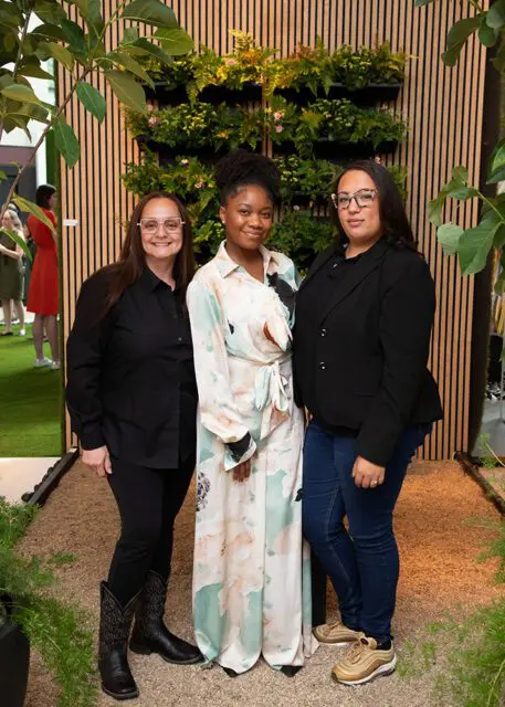 Three women posing in front of a vertical garden wall and wood slat wall.