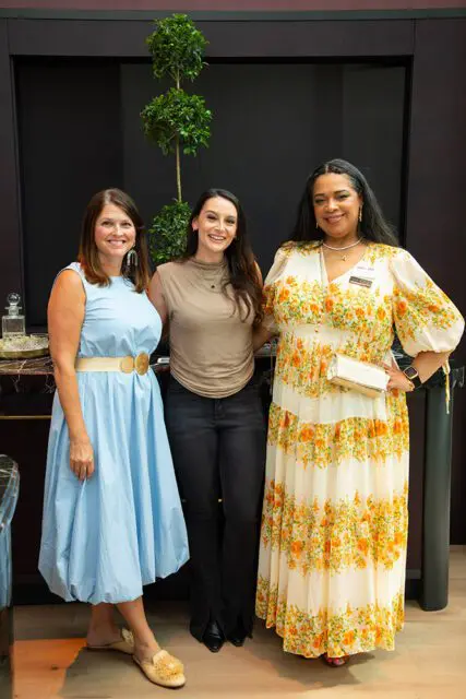 Three women standing together, one in a blue dress, one in a taupe top and black jeans, and one in