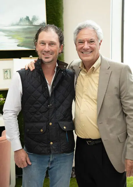 Two men smiling, one in a quilted vest and jeans, the other in a tan suit and yellow shirt.