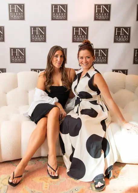 Two women sitting on a white couch in front of a wall with Harry Norman Realtors logos.