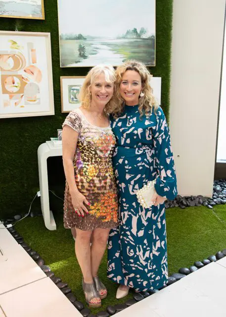 Two women smiling, one in a patterned dress and the other in a teal dress with a pearl clutch.